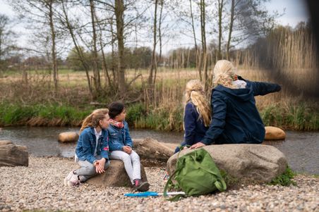 Mutter macht mit ihren Kindern Pause im Wassererlebnisbereich im Regionalpark Wedeler Au. Alle sitzen auf großen Steinen am Ufer.