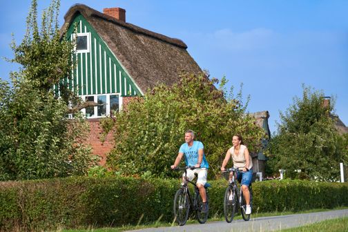 Paar ist mit dem Fahrrad unterwegs und kommt gerade an einem hübschen Reetdachhaus vorbei.