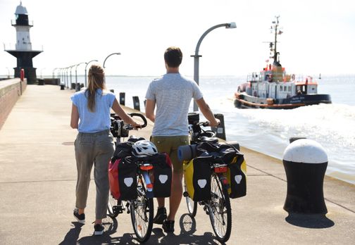 Paar ist mit dem Fahrrad in Brunsbüttel unterwegs und schiebt die Fahrräder in Richtung Mole und genießt den Ausblick auf die Elbe.