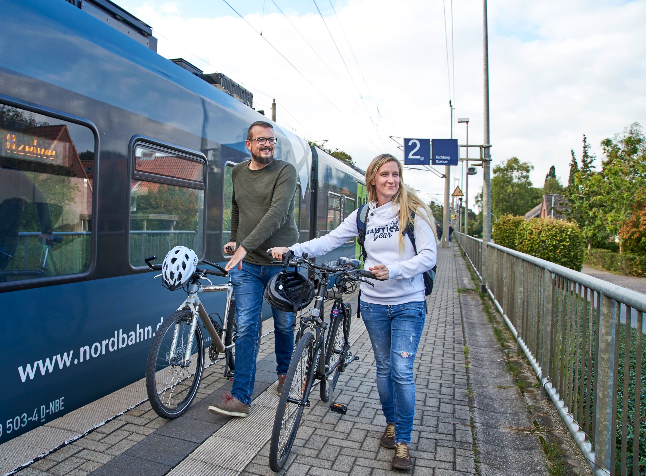 Ein Paar schiebt Fahrräder über den Bahnsteig. Im Hintergrund steht der haltende Zug.