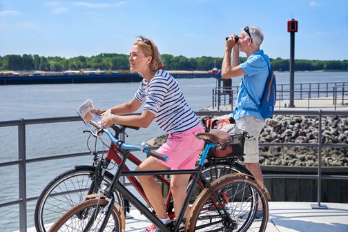 Paar ist mit dem Fahrrad in Wedel unterwegs, macht eine Pause an der Schiffsbegrüßungsanlage und genießt den Blick auf die Elbe.