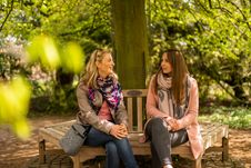 Zwei Freundinnen sitzen auf einer Bank unter einem großen Baum und genießen eine Pause im Arboretum Ellerhoop.