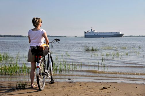 Eine Frau steht am Elbstrand neben ihrem Fahrrad und genießt den Blick aufs Wasser.