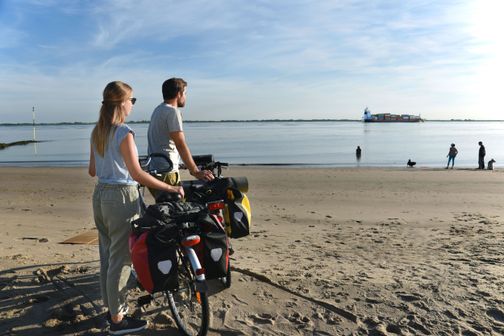 Ein Paar ist mit dem Fahrrad unterwegs und macht eine Pause am Elbstrand in Brokdorf. Beide stehen neben ihren Rädern und genießen den Blick aufs Wasser und die Schiffe.