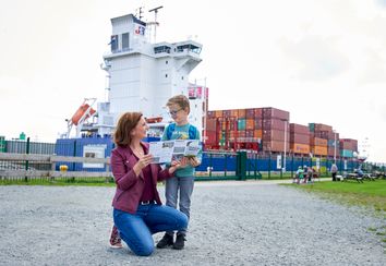 Eine Mutter besichtigt mit ihrem Sohn die Schleusenanlagen in Brunsbüttel. Im Hintergrund ist ein großes Containerschiff in der Schleuse zu sehen.