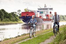 Ein Paar ist auf der NOK-Route mit dem Fahrrad unterwegs. Auf dem Kanal fährt ein großes Containerschiff.