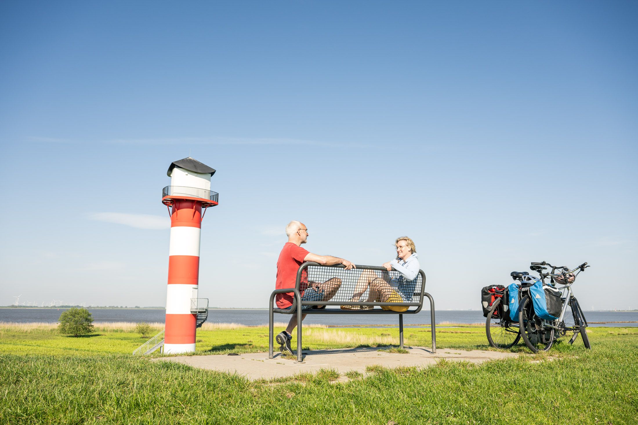 Paar ist auf dem Radfernweg Mönchsweg unterwegs und macht eine Pause auf einer Bank auf dem Elbdeich mit Blick aufs Wasser und einen Leuchtturm.