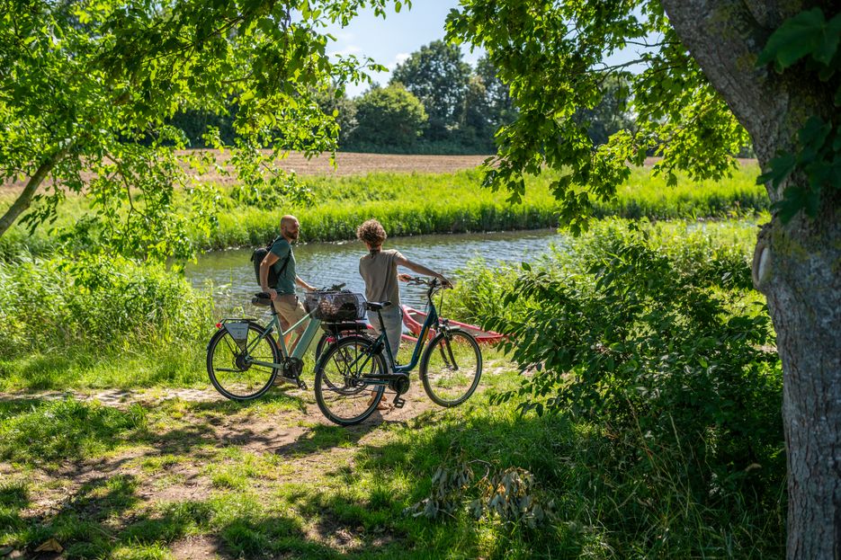 Paar steht mit dem Fahrrad am Ufer eines Flusses und schaut aufs Wasser.