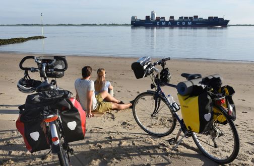 Paar ist mit dem Fahrrad auf dem Elberadweg unterwegs und genießt eine Pause am Elbstrand in Brokdorf und beobachtet die Schiffe.