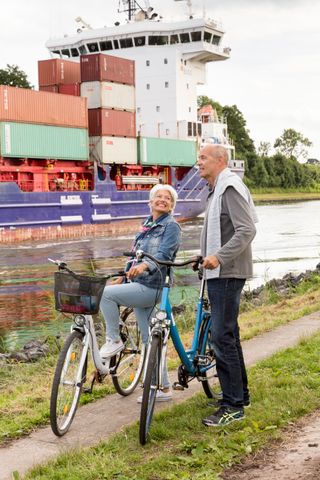 Ein Paar ist auf der NOK-Route mit dem Fahrrad unterwegs, hält gerade an und bestaunt ein großes Containerschiff.