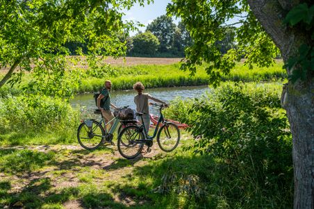 Paar steht mit dem Fahrrad am Ufer eines Flusses und schaut aufs Wasser.