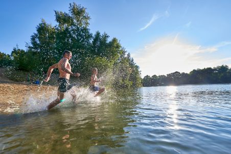 Ein Vater läuft mit seinem Sohn freudig ins Wasser. Das Wasser spritzt nach allen Seiten.