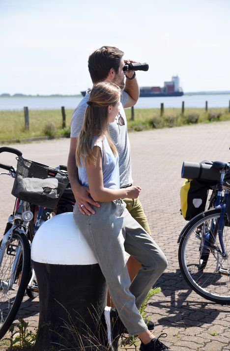 Ein paar ist mit dem Fahrrad unterwegs, macht an einer Mole in Brunsbüttel Rast und genießt den Blick auf die Elbe und die Schiffe.