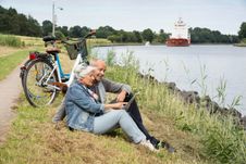 Ein Paar ist auf der NOK-Route mit dem Fahrrad unterwegs. Beide machen eine Pause und sitzen am Ufer.Auf dem Kanal fährt ein Schiff.